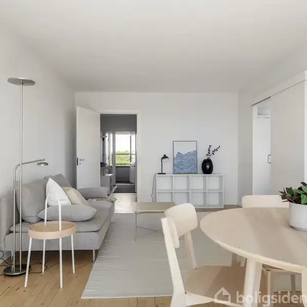 Sofa står i en lys stue med en gulvlampe ved siden af. Rundt spisebord er i forgrunden. Hylde og kunstværk ses i baggrunden.