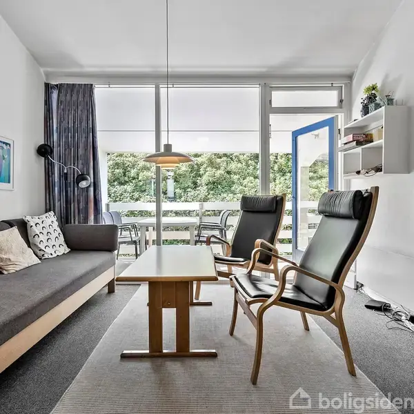 Sofa og to lænestole står i en moderne stue med tæppe og sofabord. Et tv er monteret på væggen. Store vinduer åbner op til en balkon med udsigt over grønne træer.