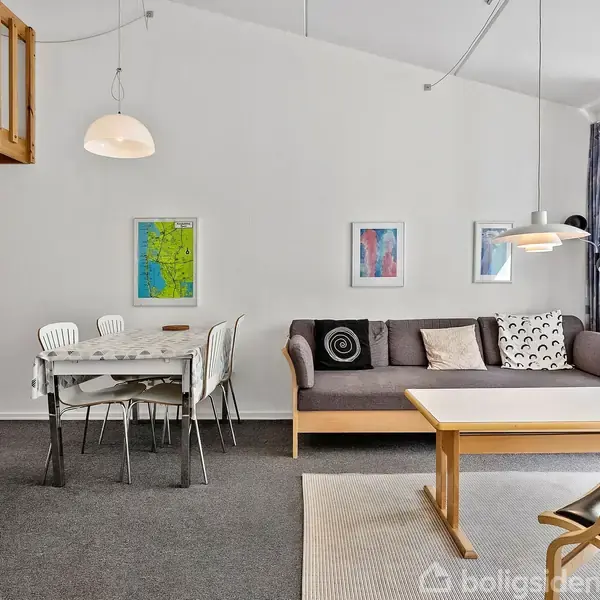 Stue med grå sofa og puder ved siden af et træ-sofabord. Ovenlys belyser rummet med spisebord og stole under vægkunst. En trappe fører opad fra køkkenområdet.