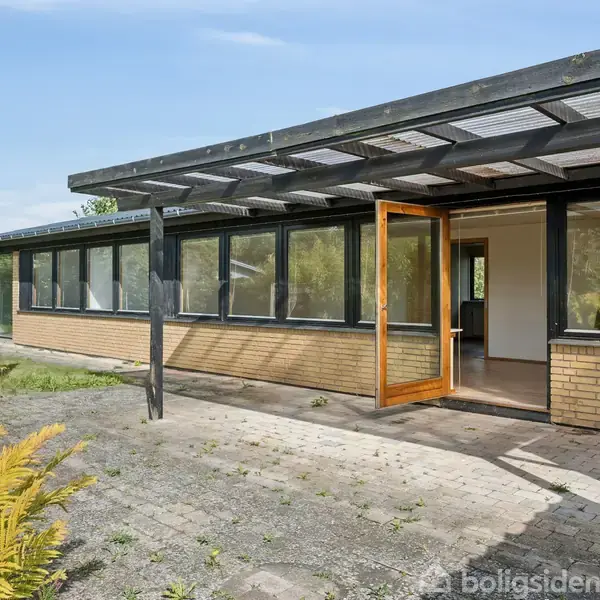 En moderne bygning med en overdækket terrasse og store vinduer. Døren er let åben, og omgivelserne viser en have med buske og en flisebelagt terrasse.