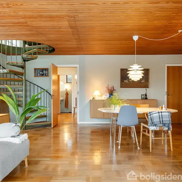 En stue med træloft og parketgulv, hvor en vindeltrappe leder opad. En grå sofa står ved siden af et lille bord med lampe, og spisebordet har stole omkring.