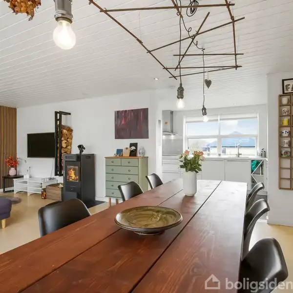 En lang, mørk spisebord med sorte stole står i et lyst, moderne stueområde med pejs. Til venstre en sofa, og bagved et stort vindue med udsigt til naturen. En plakat, en reol med kopper og en kommode dekorerer rummet.