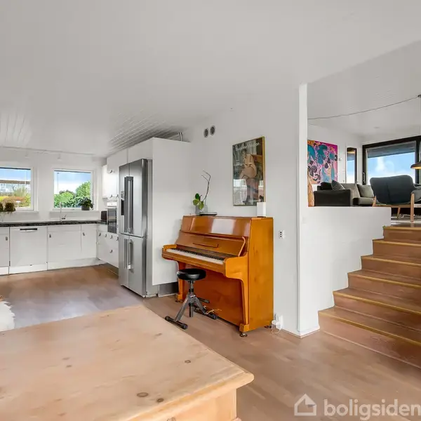 Et orange klaver står i et moderne, lyseblåt køkken med hvide elementer og et stort køleskab. Til højre fører en trappe op til en stue med store vinduer.
