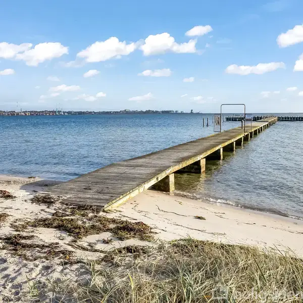 En træbro strækker sig ud i havet fra en sandstrand. Stranden er omkranset af græs og tang, under en blå himmel med spredte skyer. Ingen synlig tekst.
