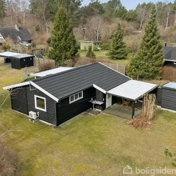 Sort sommerhus beliggende på en græsplæne med træer og buske omkring. Flere andre huse er synlige i baggrunden i et skovklædt område.