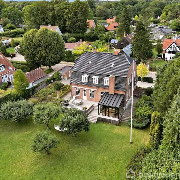 Et stort, rødt murstenshus med sort tag står på en velplejet grøn græsplæne. Huset har en udestue, der vender mod haven. Omgivet af grønne træer og naboers villaer.