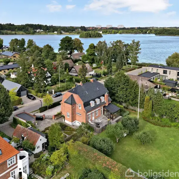 Rødt murstenshus med sort tag, beliggende i et grønt boligområde nær en stor sø. I nærheden er der andre huse, store træer og velplejede haver.