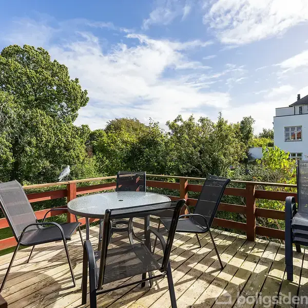 Terrassemøbler står på en træterrasse med fire stole omkring et rundt bord; omgivet af natur med frodig vegetation, og et hvidt hus med et tårn ses i baggrunden.