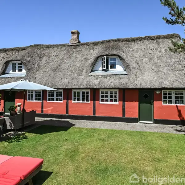 Stråtækt bindingsværkshus med røde vægge og hvide vinduer står i en velplejet have. Omkringliggende blomsterbede og havemøbler under en parasol skaber en hyggelig, landlig atmosfære.