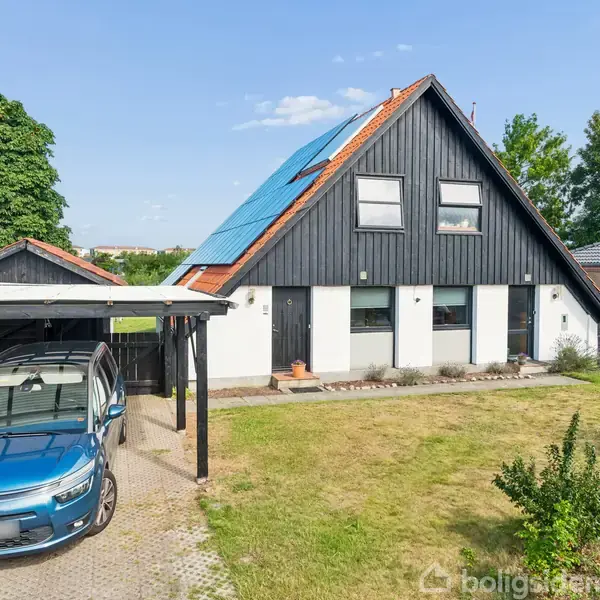 Et dansk parcelhus med sort træfacade og solpaneler på taget. En blå Citroën bil holder under en carport til venstre. Haven er grøn med enkelte buske og træer.