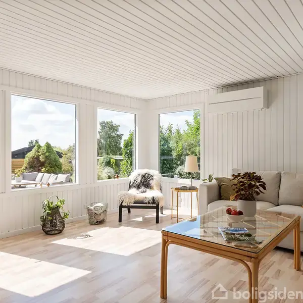 Stue med hvidt loft og vægge, lysindfald fra store vinduer, beige sofaer med farvet pude, træbord med urtepotte, lænestol, og lamper, omgivet af grønne planter udenfor.