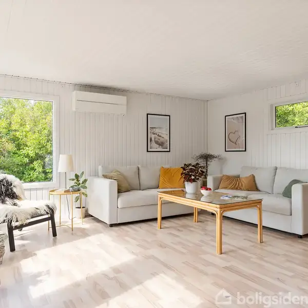 Stue med hvide vægge og trægulv, indeholdende en hvid sofa med farverige puder, et sofabord, en stol med fåreskind, og planter, omgivet af store vinduer med udsigt til naturen.