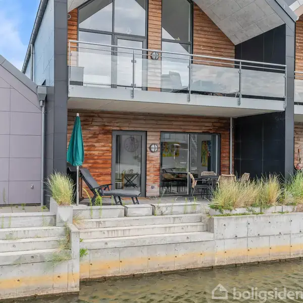 Moderne rækkehus ved vandet, med træbeklædning og glasfacade. Foran er en terrasse med havemøbler og parasol. Omgivet af trapper og betonvægge, der leder ned til kanalen.