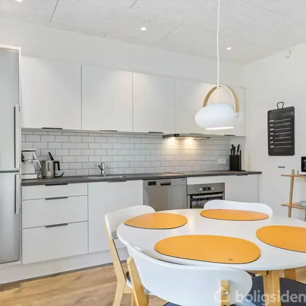 En moderne køkken med hvide skabe, køleskab, indbygget ovn, og en rund spisebord med fire stole med orange bordskånere. En tavle hænger på væggen, lys hænger over bordet.