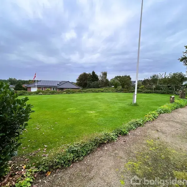 En flagstang står i en velplejet grøn græsplæne, omgivet af buske og en grussti, med et hus i baggrunden under en overskyet himmel.
