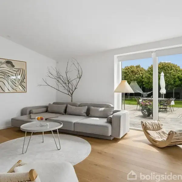 Stue med grå hjørnesofa ved et rundt sofabord, dekoreret med frugt, oplyst af en gulvlampe. Store vinduer åbner ud til terrasse og have med træer.