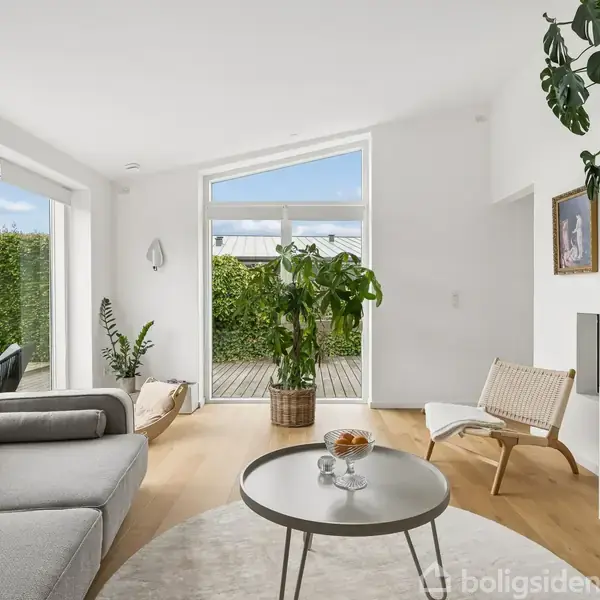 En stue med en grå sofa og lampe, placeret ved glasdøre, som leder til haven. En rund bordplade med dekorationer, omgivet af grønne planter og en bogreol fyldt med farverige bøger.