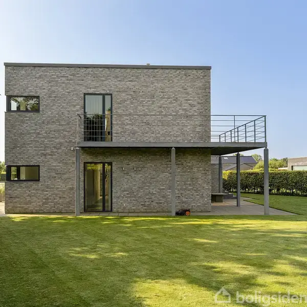 En moderne murstensbygning med fladt tag står stille på en velplejet græsplæne omgivet af hække. Sollys skinner ind på facaden, og der er en altan på første sal.