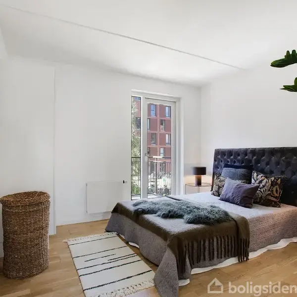 En seng med mørkt sengegavl står midt i et lyst soveværelse. En plante og kurv er i hjørnet, mens store vinduer slipper lys ind. En stribet tæppe er på gulvet.