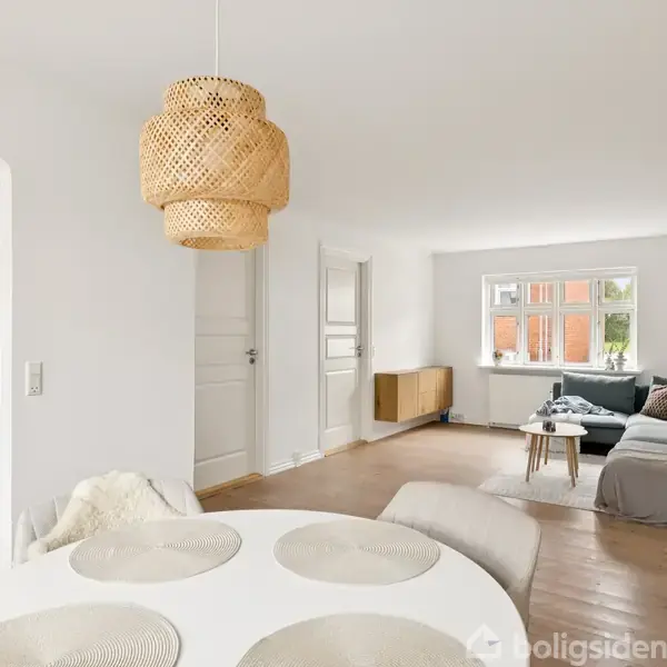 Rundt bord med dækkeservietter står i en lys stue med en sofa og sofabord. En køkkenåbning viser skabe. En flettede lampeskærm hænger fra loftet.