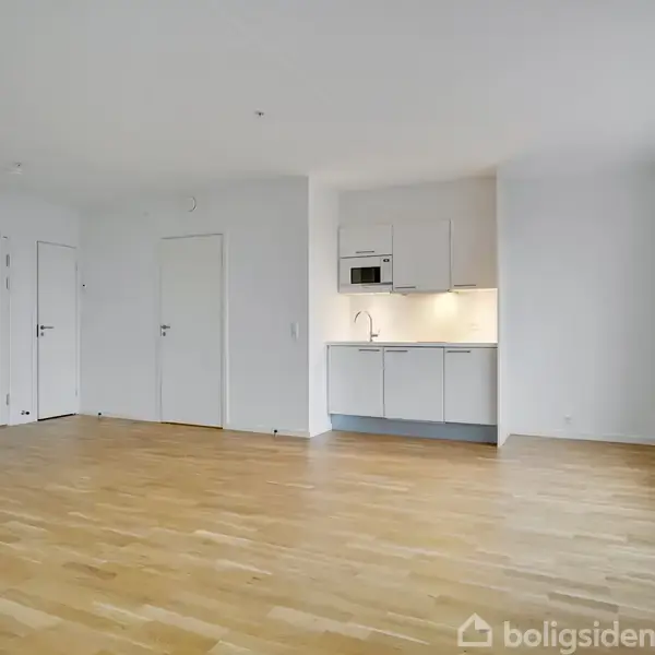 Et moderne køkken integreret i en lys stue. Køkkenet har hvide skabe, en vask, og en indbygget mikrobølgeovn. Rummet har trægulv og en stor, lysende vindueskarm.