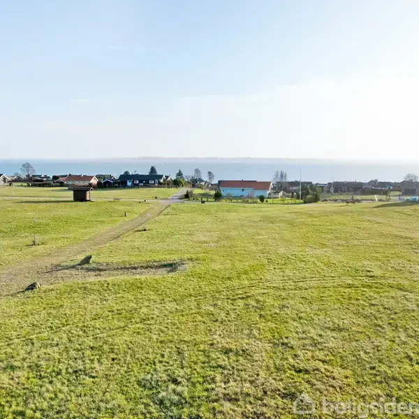 Græslandet strækker sig mod en række huse og træer i baggrunden, med en udsigt til havet under en klar himmel.