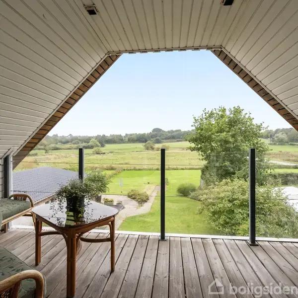 Balkon med to kurvestole og et træbord med en plante, vender ud mod en frodig have og åbent landskab med grønne marker og spredte træer i horisonten.