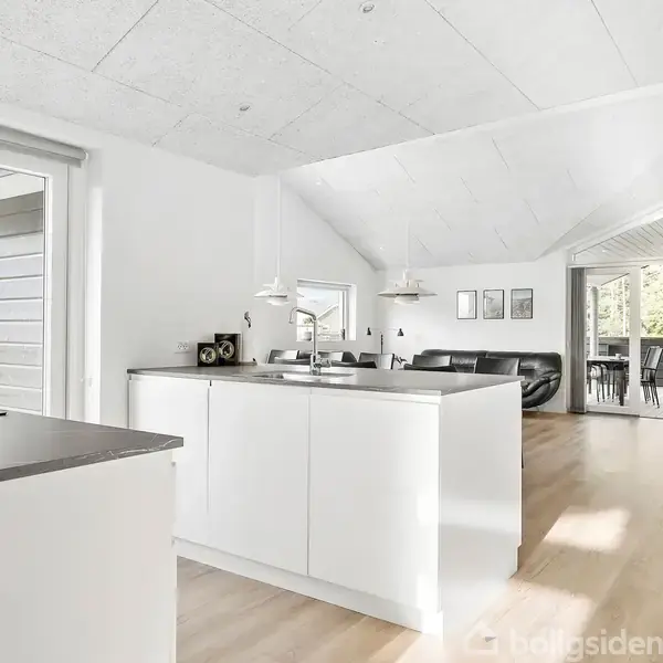Et moderne køkken med hvide skabe og mørk bordplade åbner op til en lys stue. Der er store vinduer, som viser en terrasse og grøn have udenfor.