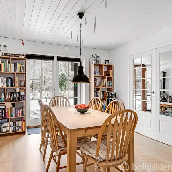 Et spisebord med seks stole står under en sort lampe. Hylder fyldt med bøger omkranser rummet. Vinduer og glasdøre giver udsigt til en sneklædt have.