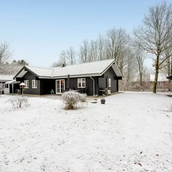 Sort træhus står stille i snedækket have omgivet af nøgne træer.