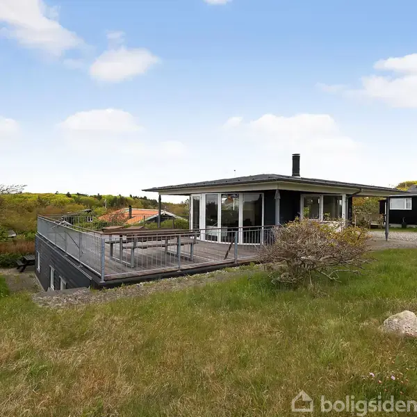 Et moderne sommerhus med store panoramavinduer og en træterrasse. Huset står på en græsbeklædt bakke, omgivet af en frodig landskab under en blå himmel med lette skyer.