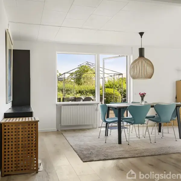 Spisebord med blå stole står på et tæppe i et lyst rum. En stor lampe hænger over bordet, og vinduer viser en frodig have udenfor.