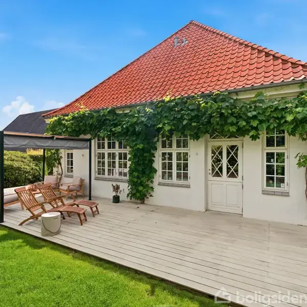 Et hvidt hus med rødt tegltag har en træterrasse med havemøbler og pergola. I krattet omkring vokser grønne planter. Græsplæner omgiver huset under en blå himmel.