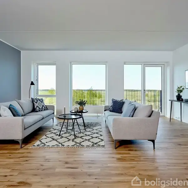 To sofaer står i en stue med parketgulve og hvidmalede vægge, under en stor glaspanel med udsigt. Der er et rundt sofabord og grønne planter i rummet.