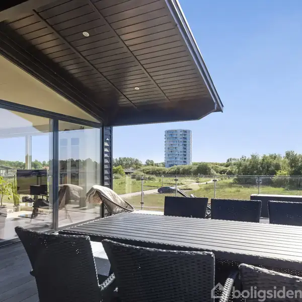 En udendørs terrasse med et sort bord og stole, under en stor tagudhæng. I baggrunden ses en græsplæne, træer og en høj bygning i det fjerne.