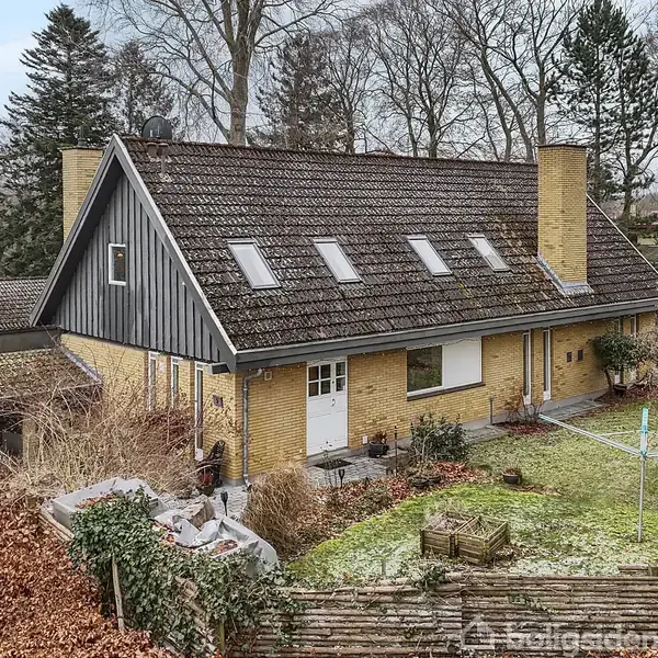 Hus står stille i en have med vinterbrune buske og træer. Det har et skrånende tag, fire ovenlysvinduer, og to skorstene. Græsplæne med tørresnor ses i nærheden.
