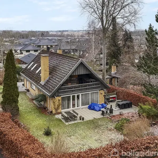 Et hus med skråt tag og stor terrasse står stille i en villakvarter, omgivet af hække og høje træer på en græsplæne.