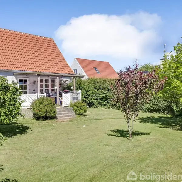Hus med rødt tegltag står stille i en solrig have med græsplæne, buske og træer. Træterrasse ved indgangen har trapper, omkranset af hække.