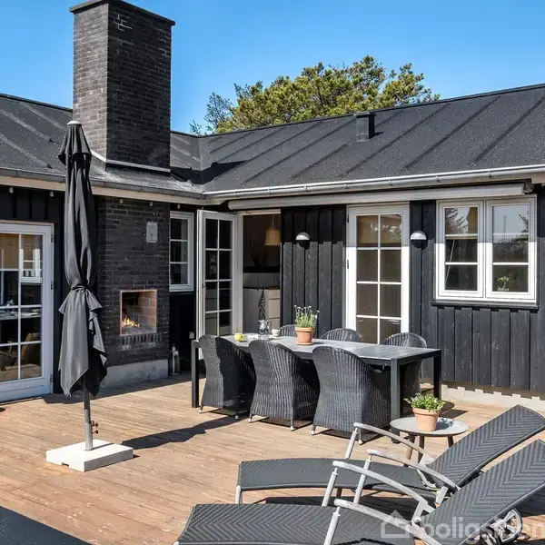 Terrasse med sortmalet hus har spisebord og stole foran, solsenge til højre og en parasol. En skorsten er synlig, og planter står rundt omkring.