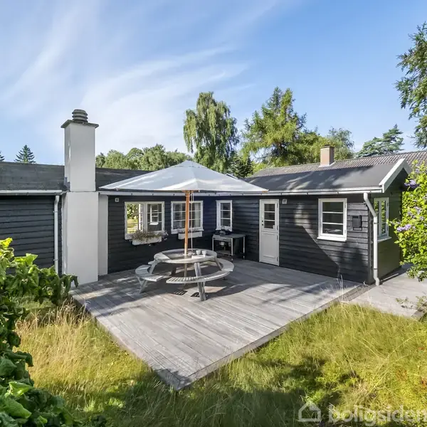 Et sortmalet hus med en terrasse, hvorpå der står et rundt bord med en parasol. Omgivet af græs og træer, ses en solrig, fredfyldt have.