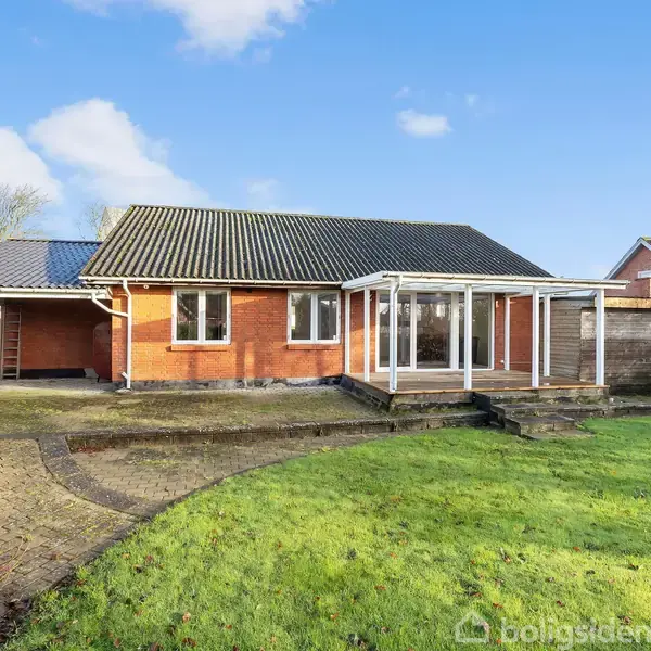 Et rødt etplanshus med mørkt tag står i en have med græsplæne og flisebelægning. Til højre er en glasveranda, og store vinduer vender mod en klar, blå himmel.