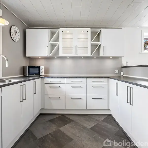 Et moderne køkken med hvide skabe arrangeret i en L-form. En mikroovn står på bordet. Vinduet viser udsigt til græsplænen udenfor. En vægur hænger på væggen.