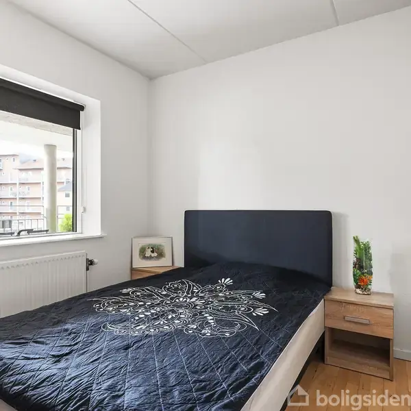 Soveværelse med en enkeltseng dækket af et blomstermønsteret sengetæppe. Ved siden af sengen står to natborde. Værelset har et vindue med udsigt til lejlighedsbygninger.