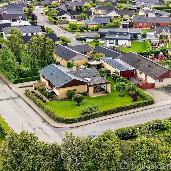 Et parcelhus med have og terrasse ligger på en hjørnegrund. Omkring er der flere villaer og grønne haver i et boligkvarter, der viser en velholdt og ordnet bebyggelse.