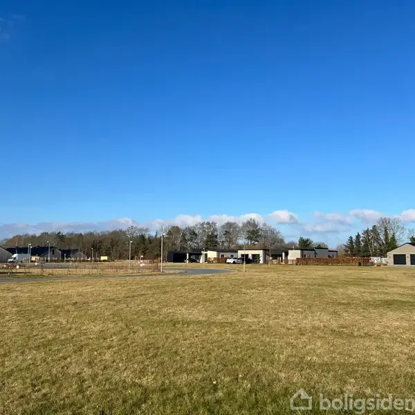 Græsmark strækker sig mod horisonten, hvor spredte, moderne huse ses under en klar, blå himmel. Træer står i baggrunden og skaber en rolig landskabsscene.