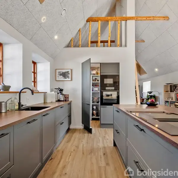 Køkken med grå skabe er åben og moderne. En ovn og mikrobølgeovn er indbygget i væggen, mens køkkenøen har en kogeplade. Træbjælker pryder det skrå loft.