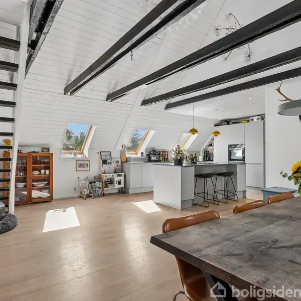 En stor moderne stue med skrå loft og træbjælker. Rummet indeholder et spisebord, stueplanter og et køkkenområde med barstole. En trappe fører til en anden etage.