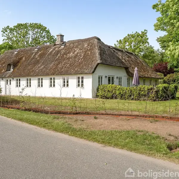 Hvid traditionel bindingsværkshus med stråtag står stille ved vejkant. Husets have er omgivet af grøn vegetation og et lille hegn, med en parasol synlig i haven.