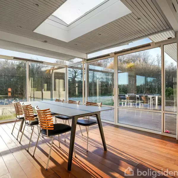 En spisebord med stole står i en lys vinterhave med glasvægge. Udsigt til en terrasse og have udenfor med træer i baggrunden. Skylight i loftet.