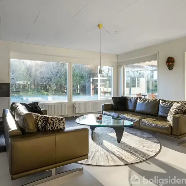 En stue med sofagruppe omkring et cirkulært glasbord. Sollys strømmer ind gennem store vinduer og afslører en have udenfor. Væggen har dekorative elementer og til venstre er en pejs.